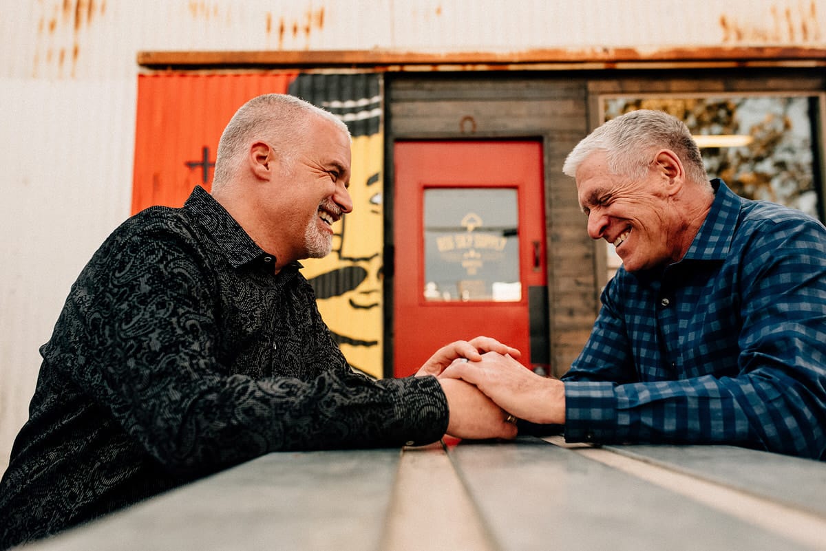 two older men with grey hair sitting across the table from each other holding hands smiling