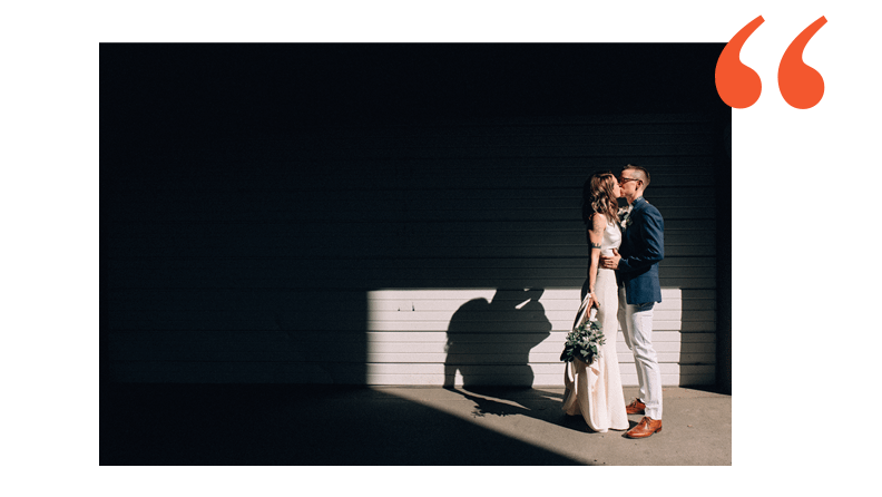 lesbian couple standing in a pocket of light in an alleyway. one is wearing a sleek off white wedding dress and the other is wearing a blue suit jacket with white pants and brown shoes.